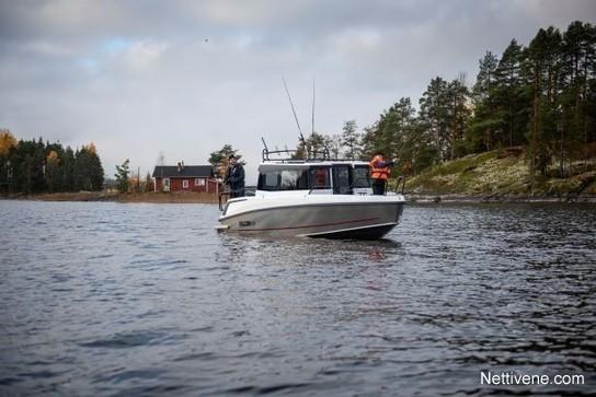 A fishing boat is on a calm lake with two people onboard, trees and a red cabin visible on the shore.